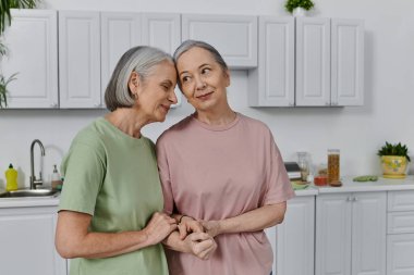 Two mature women, a lesbian couple, stand together in their modern kitchen, showcasing their love and connection. clipart