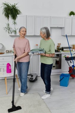 Two mature women, a lesbian couple, clean their modern apartment together. One woman vacuums while the other holds folded laundry. clipart