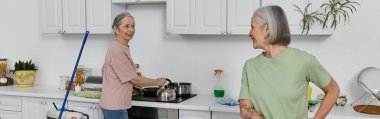 Two mature women, a lesbian couple, clean their modern apartment together. One woman washes dishes while the other watches. clipart