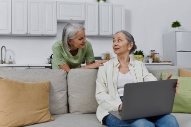 Lezbiyen bir çift, modern evlerindeki koltukta dizüstü bilgisayar kullanarak rahatlıyor.