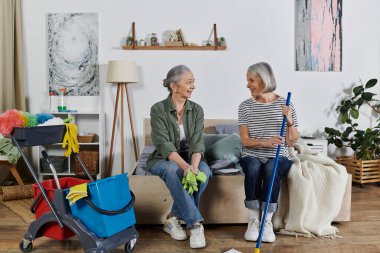 Gündelik giyinmiş iki olgun lezbiyen kadın modern dairelerini temizliyor. Biri eldiven tutarak yatağa oturur, diğeri paspas tutar..