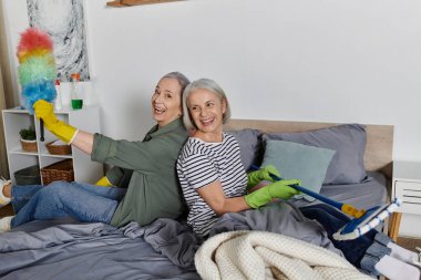 Gündelik giysiler içinde iki olgun lezbiyen kadın birlikte modern dairelerini temizlerken gülümsüyor ve eğleniyorlar..