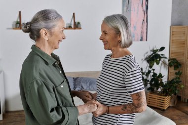 A mature lesbian couple smiles at each other in their modern apartment. clipart