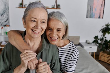 İki olgun kadın, modern bir dairede birbirine sarılıp gülüyor ve sevgi dolu bir ilişki sergiliyorlar..