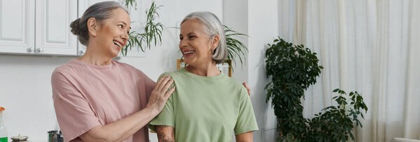 Две пожилые лесбиянки смеются в современной квартире, одна заканчивает работу по дому.