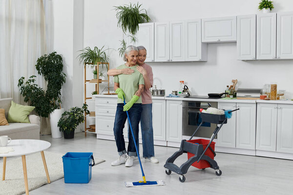 A lesbian couple, both with gray hair, cleans their modern apartment together. The woman on the left is mopping the floor.