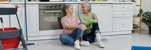 Две зрелые женщины, пара лесбиянок, отдыхают от уборки современной кухни, сидят на полу и говорят:.