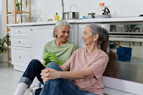A mature lesbian couple, dressed casually, are cleaning their modern apartment together, taking a break to share a laugh.