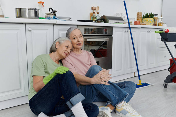 Взрослая лесбийская пара отдыхает от уборки своей современной кухни, когда одна женщина кладет голову на плечо.