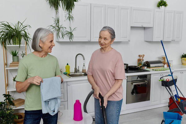 Two mature lesbians are cleaning their modern kitchen together, one holding a cleaning cloth and the other holding a mop.