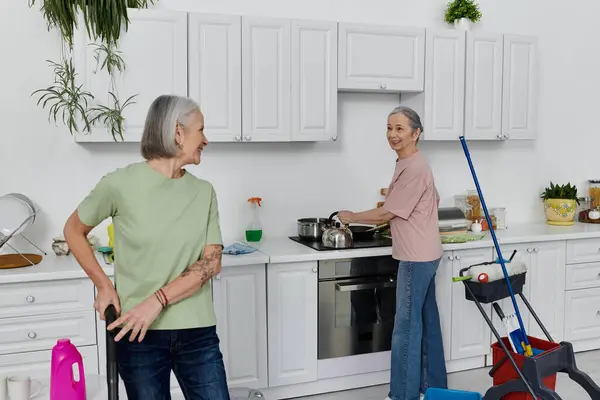 stock image A lesbian couple cleans their modern kitchen together. One woman holds a mop while the other washes dishes.