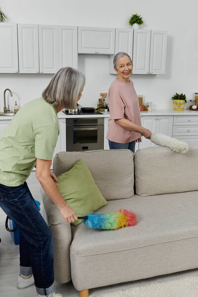 An older lesbian couple cleans their modern apartment together, one woman using a duster on the couch.