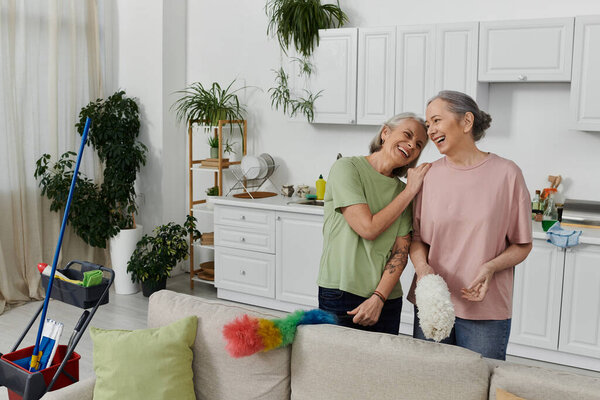 A lesbian couple laughs together while cleaning their modern apartment.