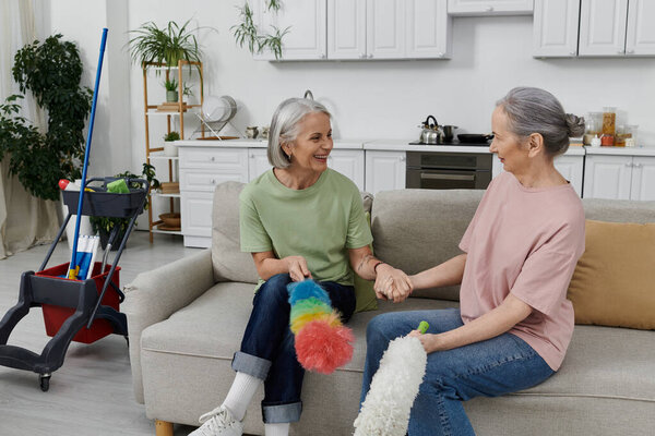 A mature lesbian couple smiles and chats while cleaning their modern apartment together.