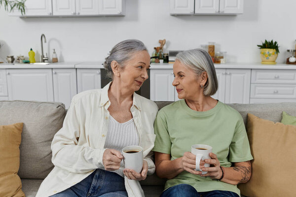 Two mature lesbian women relax on a couch in their modern apartment, enjoying coffee after a cleaning session.
