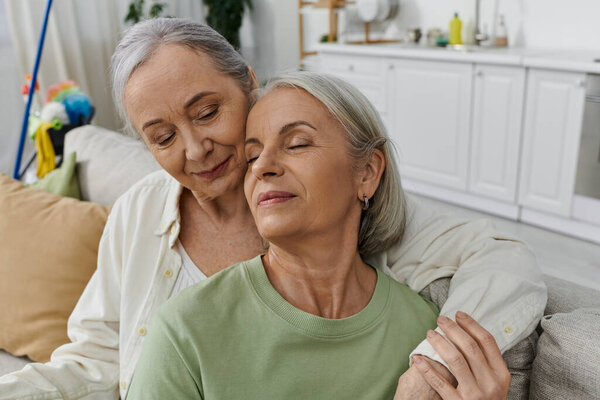 A mature lesbian couple relaxes on a couch after cleaning their modern apartment, embracing love and contentment.