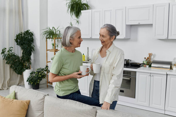 Two mature women, a lesbian couple, relax on a couch in their modern apartment after cleaning, enjoying coffee.