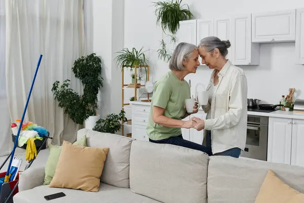 stock image A mature lesbian couple rests after cleaning their modern apartment, holding mugs and sharing a loving moment.