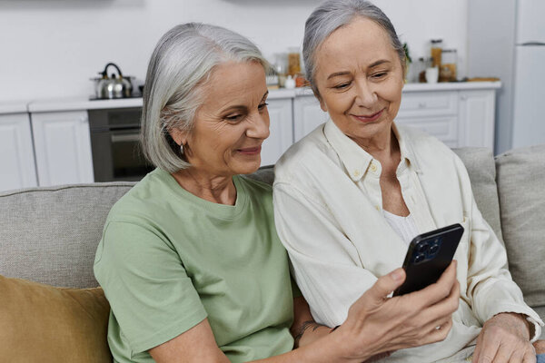 Лесбийская пара, отдыхающая на диване в своей современной квартире, улыбается и смотрит в телефон после окончания уборки.