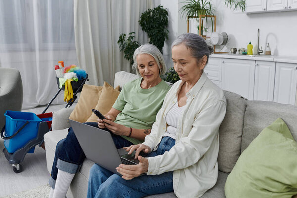 Две зрелые лесбиянки отдыхают на диване после уборки в своей современной квартире, одна женщина пользуется ноутбуком, а другая телефоном.