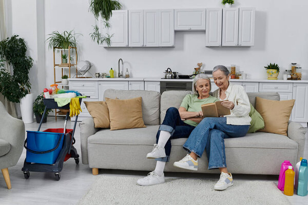 Две зрелые лесбиянки отдыхают на диване в своей современной квартире после уборки, на заднем плане видны чистящие средства.