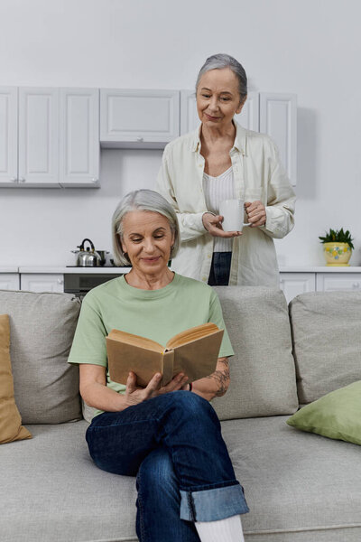 Пара отдыхает на современном диване после уборки. Один читает, а другой наслаждается чаем в своей квартире.