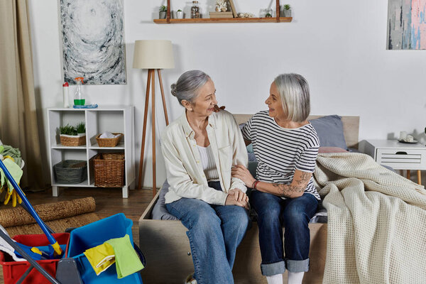 Two mature lesbian women rest on a bed in their modern apartment after cleaning.