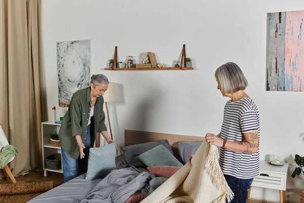 A mature lesbian couple makes their bed in their modern apartment.