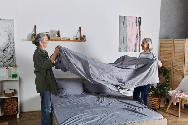 Two mature lesbian women make their bed together in a modern apartment.