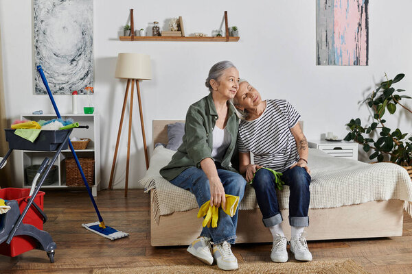 Две зрелые лесбиянки отдыхают на своей кровати в современной квартире после уборки, демонстрируя любовные отношения и уютный дом.