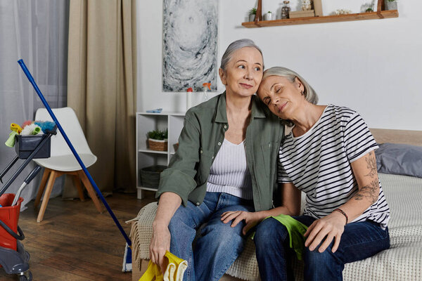 Two mature lesbian women take a break from cleaning their modern apartment, resting together on the couch.