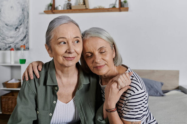 Two mature lesbian women in casual attire sit together in a modern apartment, sharing a tender moment.