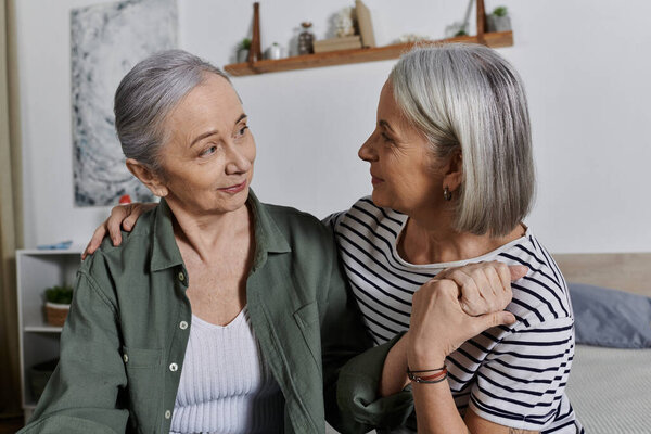 Two mature lesbian women in casual attire share a tender moment together in their modern apartment.