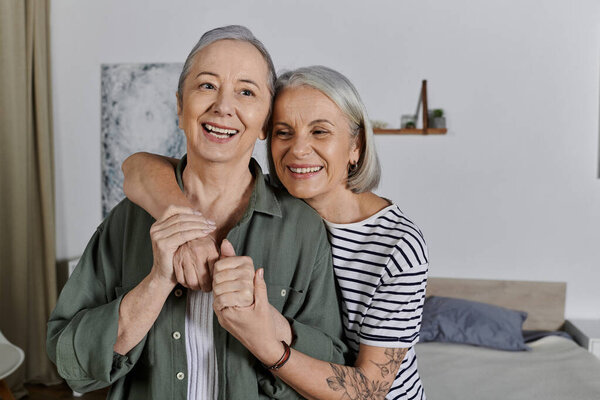 A mature lesbian couple embraces in their modern apartment. They are both smiling happily and look very much in love.