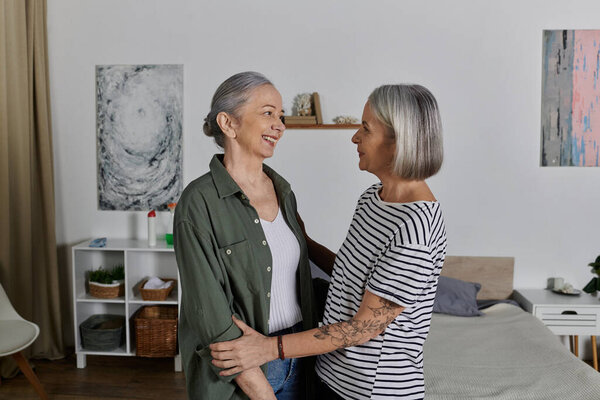 Two mature women, a lesbian couple, embrace each other in a modern apartment, their smiles radiating happiness and love.