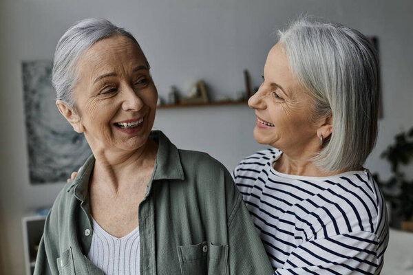 Two older women, a lesbian couple, joyfully embrace in a modern apartment. Both have gray hair and big smiles.