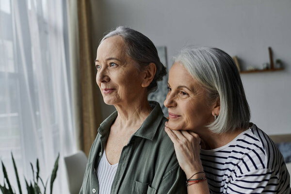 Две зрелые женщины обнимаются в современной квартире, глядя в окно с чувством удовлетворения.