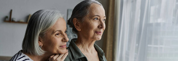 Две женщины, руки переплетаются, разделяют любящий взгляд в их современной квартире, глядя в соседнее окно.