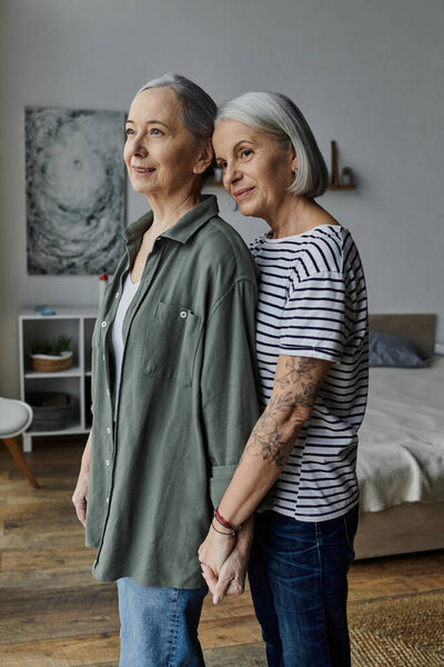 A mature lesbian couple stand in their apartment, arms wrapped around each other, sharing a tender moment.