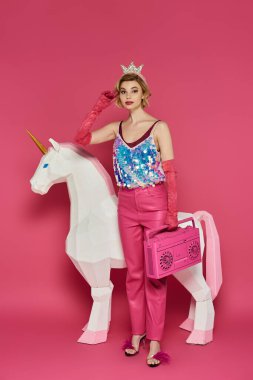 Pembe elbiseli ve taçlı bir kadın beyaz bir tek boynuzlu at ve pembe bir teyple poz veriyor..