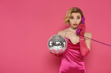 Pembe elbiseli ve uzun eldivenli bir kadın klasik bir telefonla konuşurken bir disko topu tutuyor..