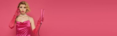 Pembe elbiseli ve eldivenli bir kadın pembe saç düzleştiricisini pembe arka planda tutuyor..