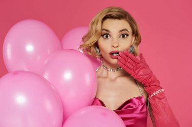 Pembe elbiseli ve eldivenli şaşırmış bir kadın pembe balonların arasında duruyor..