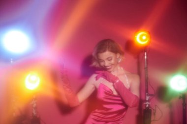 Canlı pembe elbiseli bir kadın renkli sahne ışıkları altında poz verir..