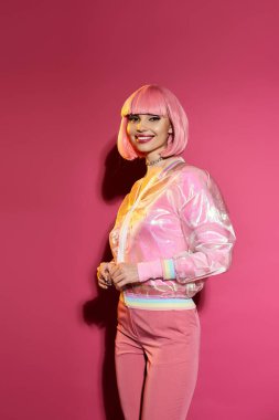 Pembe saçlı ve parlak pembe elbiseli bir kadın pembe bir arka plana güvenerek gülümsüyor..