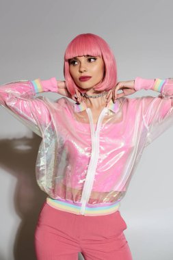 Pembe saçlı ve şeffaf ceketli bir kadın beyaz bir fonda poz veriyor..
