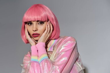 Pembe saçlı ve parlak pembe ceketli genç bir kadın gri bir arkaplanda poz veriyor..