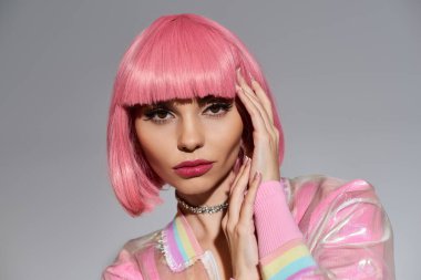 Pembe saçlı ve parlak pembe ceketli genç bir kadın gri bir arkaplanda poz veriyor..