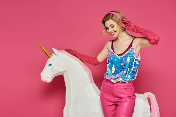 Женщина в розовом наряде улыбается, слушая музыку в наушниках, позируя рядом с белым единорогом.