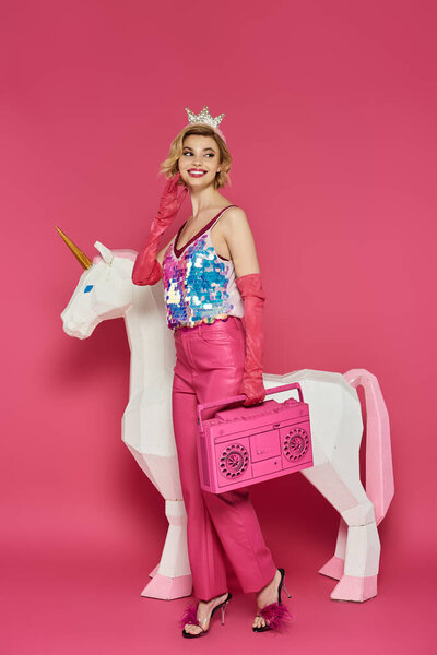 Женщина в розовом наряде и корона позирует с белым единорогом и розовым бумбоксом.